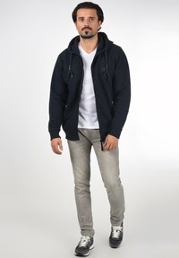 Hoodie zippé marine avec cordons, t-shirt blanc en dessous, jean gris clair et sneakers marines. Fond neutre avec un éclairage doux.