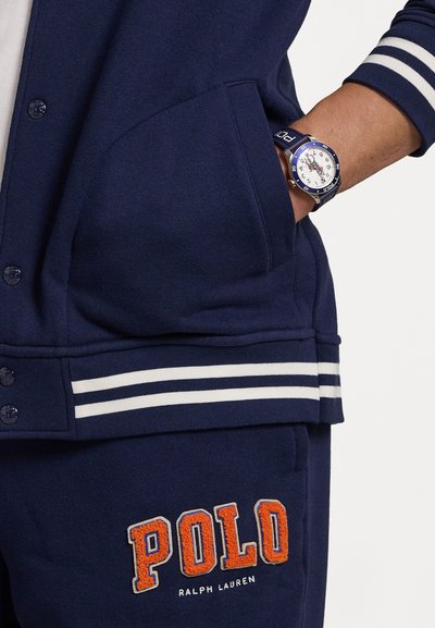 Navy sweatshirt with white striped accents and buttons, featuring an orange "POLO" embroidered logo on the pocket. Blue watch with white face visible.