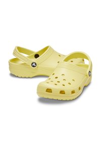 Sabots moulés jaunes avec un bout arrondi, des trous de ventilation, une sangle au talon et une semelle texturée. Présente un logo circulaire sur le côté.