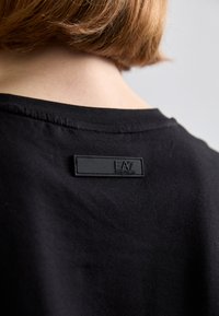 Musta puuvillapaita, jossa on sileä tekstuuri ja musta nahkainen tuotemerkkilappu takana kohokuvioidulla logolla.