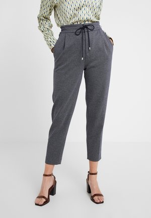 Kobieta ubrana w szare, zwężane spodnie z wysokim stanem i sznurkiem, wzorzystą koszulę z długim rękawem oraz ciemnobrązowe sandały na słupku.