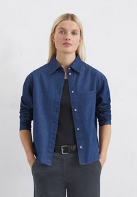 Dunkelblauses Denimhemd mit spitzem Kragen, Knopfleiste und einer einzelnen Brusttasche. Die langen Ärmel sind bis zu den Ellbogen hochgerollt.