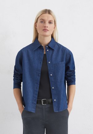 Dunkelblauses Denimhemd mit spitzem Kragen, Knopfleiste und einer einzelnen Brusttasche. Die langen Ärmel sind bis zu den Ellbogen hochgerollt.