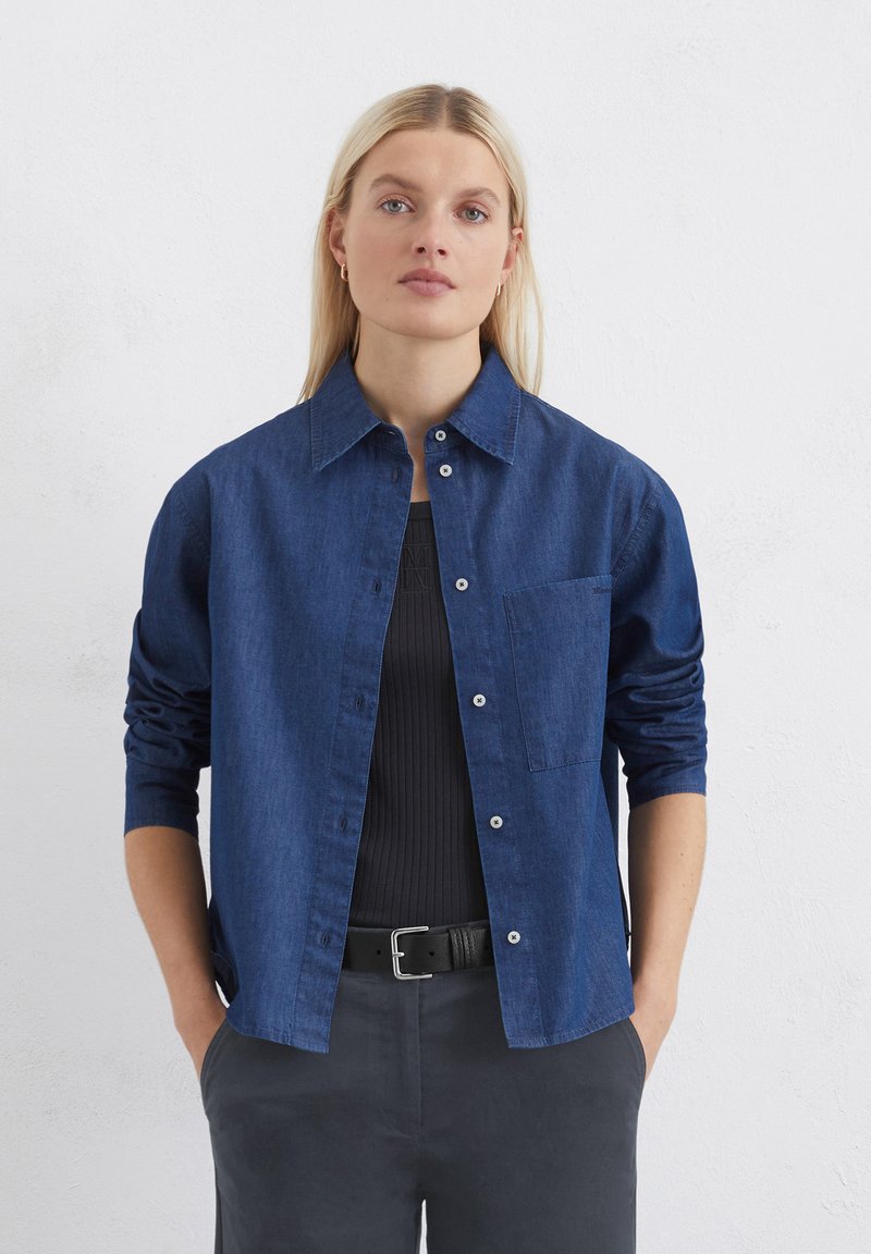 Dunkelblauses Denimhemd mit spitzem Kragen, Knopfleiste und einer einzelnen Brusttasche. Die langen Ärmel sind bis zu den Ellbogen hochgerollt.