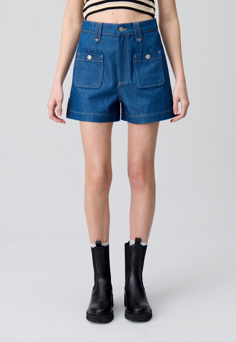 Pantalones cortos de mezclilla en azul oscuro con dos bolsillos frontales, costuras visibles y una cintura alta. Combinados con botas negras de tobillo con lados elásticos.