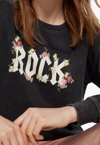 Černá mikina s květinovými akcenty kolem slova "ROCK" napsaného tučnými bílými písmeny. Měkká textilie, uvolněný design.