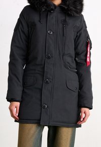 Svart parka-jacka med huva kantad med päls, dragkedja och knappstängning, två framsidor fickor och märke på ärmen. Mjuk tygkänsla.