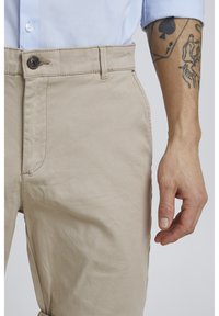 Pantalones de algodón beige con delantero plano, que presentan un cierre de botón y costuras visibles, mostrados con una mano posicionada cerca de la cintura.