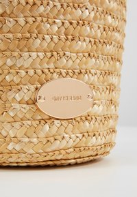 Cestino di paglia intrecciato con forma cilindrica, colore beige chiaro e una placca ovale dorata lucida con la scritta "RIVER ISLAND."