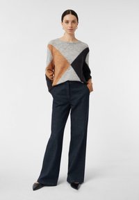 Eine Frau steht mit den Händen in den Taschen, trägt einen grau-schwarz-braunen Color-Block-Pullover, dunkle weite Hosen und schwarze spitze Schuhe.
