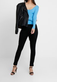 Niebieski prążkowany top z długim rękawem, czarne obcisłe jeansy i czarne sznurowane szpilki. Modelka trzyma czarną skórzaną kurtkę motocyklową.