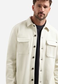 Witte gemêleerde shirtjas met een coller neckline, twee borstzakken en donkere knoopsluitingen over een zwart T-shirt eronder.