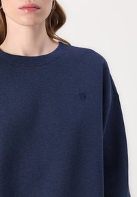 Marineblå bomullssweatshirt med rund hals, senket skuldre og brodert logo på brystet. Myk tekstur og avslappet passform.