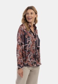 Blusa a maniche lunghe con stampa paisley in tonalità di rosso, blu navy e crema. Il tessuto è leggero con colletto a bottoni e dettagli arricciati.