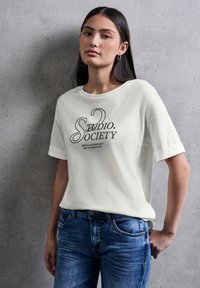 Weißes Kurzarm-T-Shirt mit dem grafischen Schriftzug "Studio Society" in dunkler Tinte, kombiniert mit blauen Jeans, vor einem grauen Hintergrund.