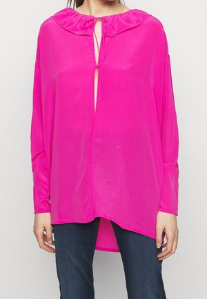 Kobieta ubrana w jasnoróżową luźną bluzkę z dekoltem w kształcie klucza, przewiązaną cienkimi sznurkami, w połączeniu z ciemnoniebieskimi spodniami.