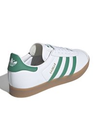 Bílé tenisky se zelenými pruhy a brandingem, vybavené texturovanou gumovou podešví a polstrovaným límcem pro pohodlí. Oceněné designové prvky Gazelle.