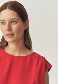 Sarkana blūze ar apaļu dekoltē un īsām, strukturētām piedurknēm. Gluda auduma virsma ar vieglu spīdumu. Modeļa mati ir taisni un brūni.
