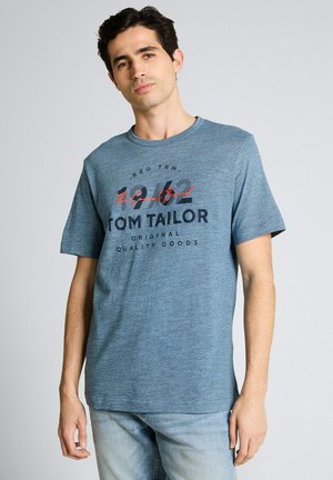 Man die een lichtblauw T-shirt draagt met de tekst "Tom Tailor Original Quality Goods", gecombineerd met lichtblauwe spijkerbroek, staand tegen een grijze achtergrond.