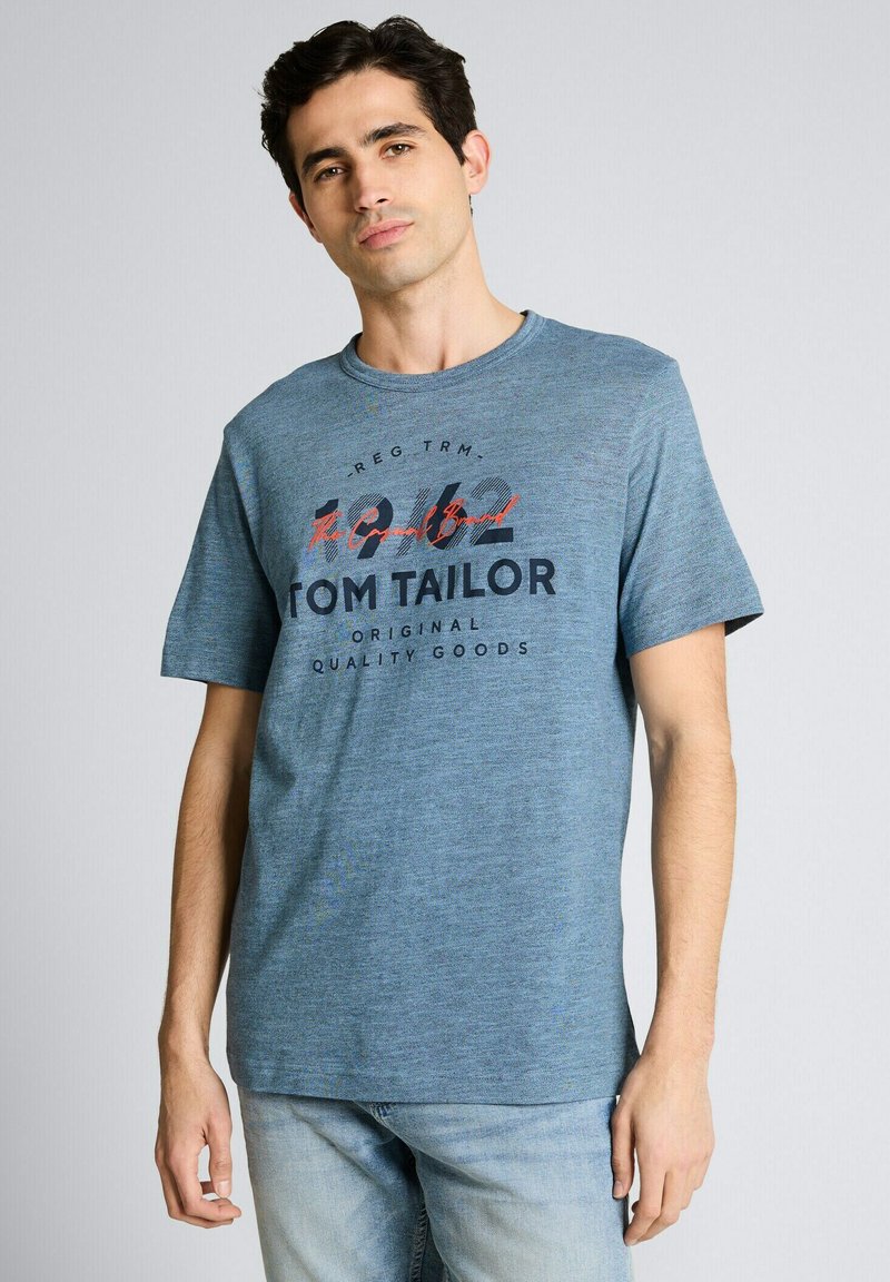 Mann trägt hellblaues T-Shirt mit Aufdruck "Tom Tailor Original Quality Goods" und hellblaue Jeans, steht vor grauem Hintergrund.