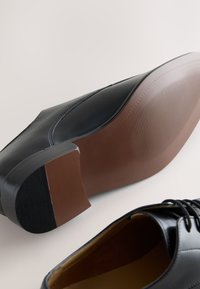 Chaussures habillées en cuir noir avec semelles marron, l'une montrant la semelle, l'autre vue de dessus avec des lacets visibles.