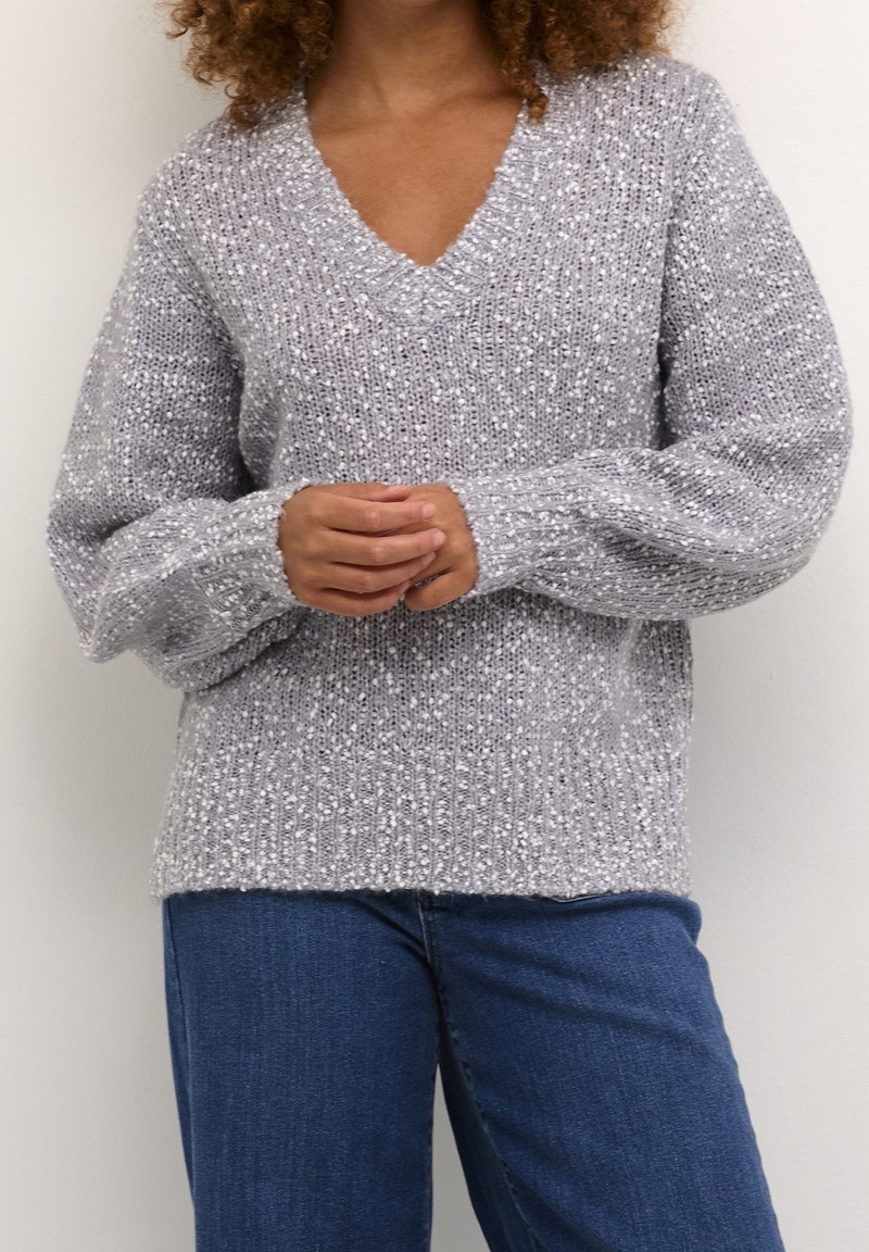Pull gris en tricot avec col en V, manches longues et motif texturé ; associé à un jean bleu, offrant une coupe décontractée et une apparence douce.