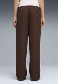 Άτομο που στέκεται με την πλάτη προς τα εμπρός, φοράει χαλαρά σκούρα καφέ παντελόνια με διακριτικό μοτίβο και λευκές πλαϊνές ρίγες, λευκό επάνω μέρος, μπροστά σε απλό φόντο.