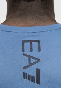 Zadnji pogled na osebo, ki nosi svetlo modro srajco z izrazitim črnim navpičnim logotipom EA in črtastim motivom pod izrezom na vratu.