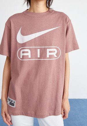 Osoba ubrana w fioletowo-różowy t-shirt Nike Air z białym logo i napisem, zestawiony z białymi prążkowanymi spodniami na jednolitym tle.