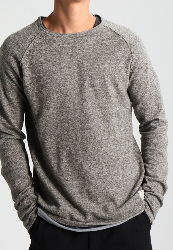 Homme portant un pull gris à col rond et manches longues, les mains dans les poches, sur un fond clair uni.