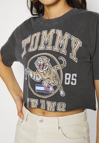 Camiseta recortada gris oscuro con un gran gráfico de un tigre rugiente, texto "TOMMY" en beige, y un pequeño logo con acentos en azul y rojo.