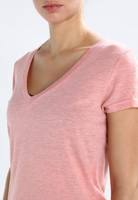 Světle růžové tričko s V-výstřihem a krátkými rukávy, vyrobené z měkké, texturované látky, s jemnými tečkami po celé ploše.