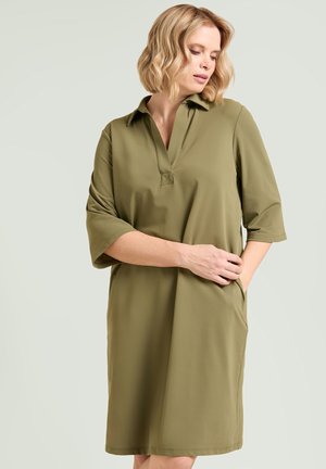 Blondynka ubrana w oliwkową, sięgającą kolan sukienkę z półdługimi rękawami i dekoltem w serek, pozująca z jedną ręką w kieszeni i patrząca w dół.
