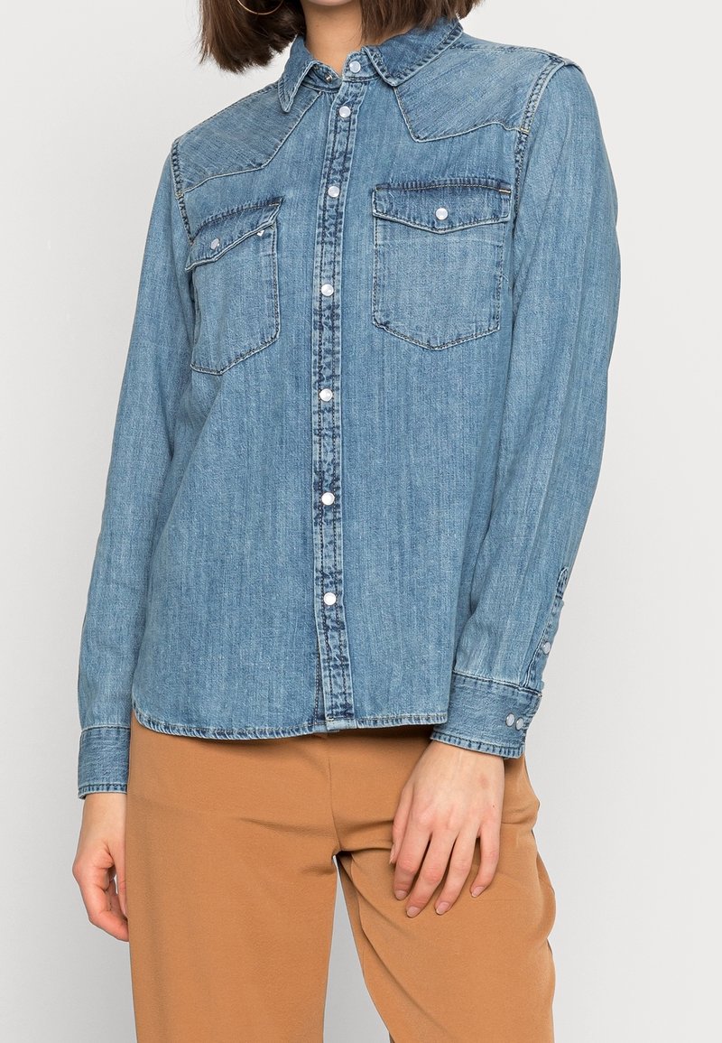 Femme portant une chemise en jean bleu clair boutonnée avec des poches poitrine à rabat et un pantalon couleur chameau, posant avec une main reposant sur la cuisse.