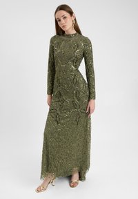 Abito da sera lungo color verde oliva con maniche lunghe, decorato con paillettes e disegni intricati, con orlo trasparente. Indossato con tacchi dorati.