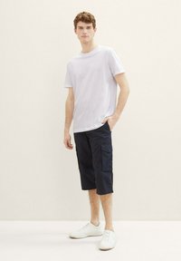 Weißes kurzärmeliges T-Shirt, marineblaue Cargo-Shorts mit seitlichen Taschen und weiße Sneaker, stehend vor einem hellen neutralen Hintergrund.