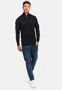 Sudadera negra con cremallera hecha de un material de punto suave, combinada con vaqueros azul oscuro y zapatillas blancas. El diseño presenta un cuello alto.