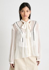 Durchsichtige weiße Bluse mit Rüschensäumen, einem Bindekragen, vertikalen Falten und schwarzen Akzenten. Spitzenverzierungen entlang der Knopfleiste sorgen für Textur.