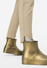 Zlaté voděodolné kotníkové boty s hladkým kovovým povrchem, s nízkou klínovou podrážkou a zipem na zadní straně, v kombinaci s béžovými kalhotami.