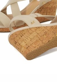 Beige leren sleehakken met kruisbanden, kurken sleehak en een gestructureerde rubberen zool. Gouden logo-accent op de band.