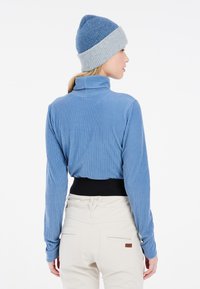 Blaues, geripptes Langarmoberteil mit hohem Kragen, kombiniert mit einer grauen Strickmütze. Weiße Hose mit einem schwarzen Bund und Gesäßtaschen.
