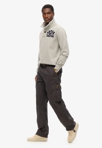 Grijze fleece trui met een hoge kraag en marineblauwe tekst, gecombineerd met donkere cargobroeken met zijzakken en een logo patch. Crème sneakers.