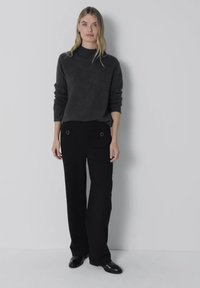 Femme debout contre un mur uni, portant un pull gris foncé, un pantalon noir à jambes larges avec des détails de boucles, et des chaussures noires.