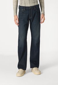 Temno modri jeans z ohlapnim krojem, ki imajo vidno šivanje, stranske žepe in zadrgo. V kombinaciji z svetlimi supergami.