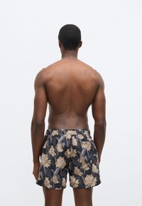 Kvetinové plavkové šortky s čiernym pozadím, zdobené veľkými béžovými kvetmi s jemným leskom. Elastický pás a detail s vreckom vzadu.