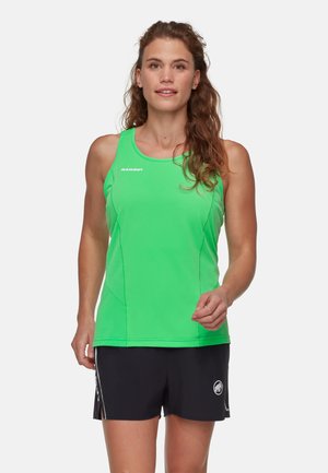 Kobieta o długich, kręconych włosach, ubrana w jasną zieloną, bezrękawnikową sportową bluzkę i czarne spodenki, stojąca zwrócona przodem na białym tle.