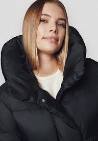 Czarna kurtka puchowa z wysokim kołnierzem, z zamkiem błyskawicznym i błyszczącymi dodatkami. Materiał wydaje się gładki i pikowany, co nadaje jej wypukły wygląd.