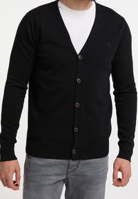 Cardigan nero lavorato a maglia con scollo a V, sei bottoni marroni e polsini e orlo a costine. Indossato sopra una maglietta bianca e abbinato a jeans grigi.