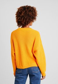 Pomarańczowy sweter dzianinowy z prążkowaną teksturą, szerokimi rękawami i skróconym fasonem. Noszony na niebieskich dżinsach, eksponującym wzór na plecach swetra.