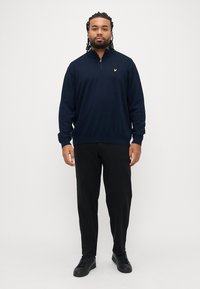 Felpa blu navy con zip realizzata in un tessuto morbido, con maniche lunghe e polsini a coste, caratterizzata da un piccolo logo dorato sul petto. Indossata con pantaloni neri.